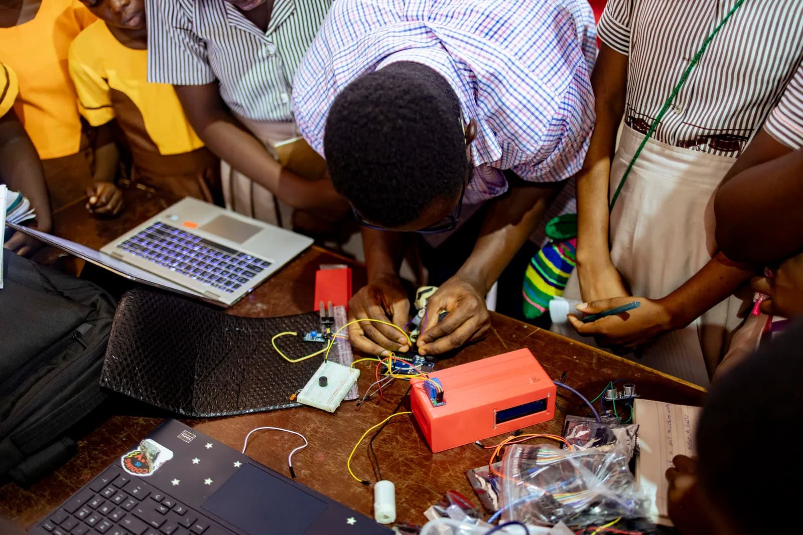 Students gathered around a hands-on electronics demonstration with a laptop, circuit boards, and sensors.